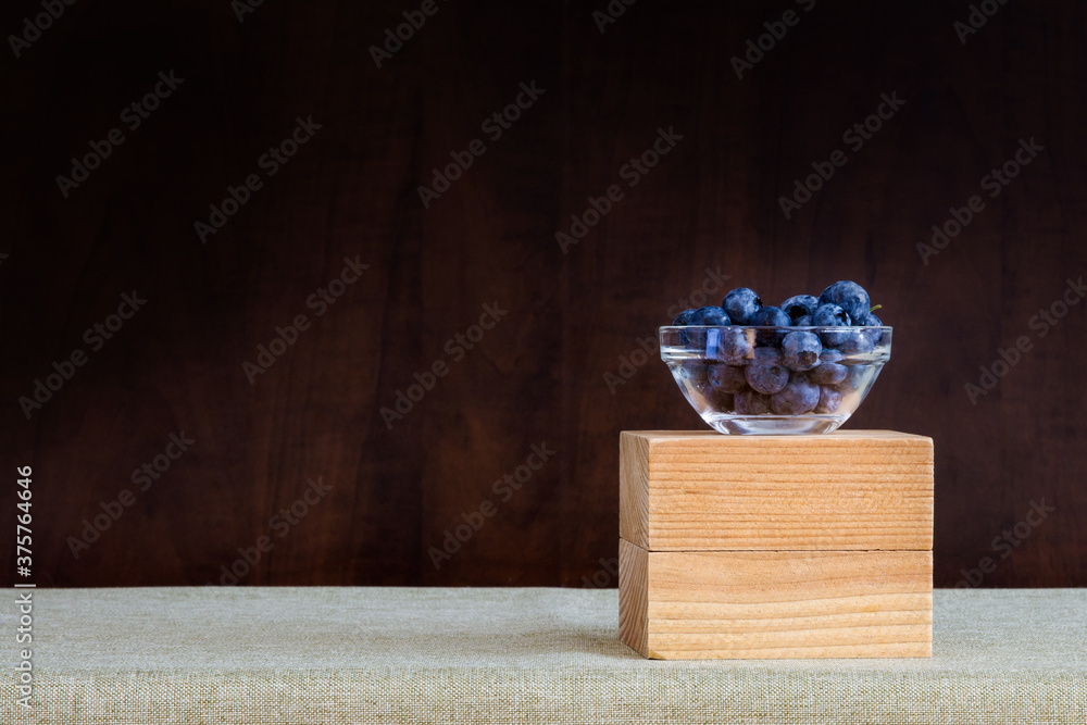 Fototapeta premium Small glass bowl of blueberries on a table with dark wood background behind; blueberries on a table covered with green table cloth