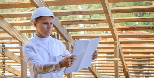 Foreman holding drawings, looking the project of wooden house, working on a construction site. Young attractive engineer in a white helmet checks the roof frame. Concept career architect. Copy space.