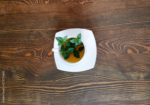 Top view of a cup with tea on a saucer. The table is made of natural wood with a pronounced texture. Aromatic herbal drink. Sprigs of fresh mint inside the mug. White tea ware.