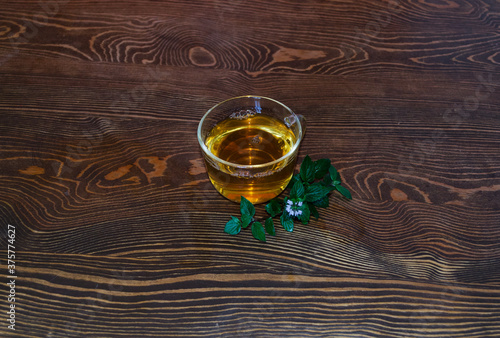 A cup of herbal tea is in the center of the table. Wooden textured background. A transparent glass mug is filled with a hot drink. Green sprigs and mint leaves lie nearby. Tranquility concept.
