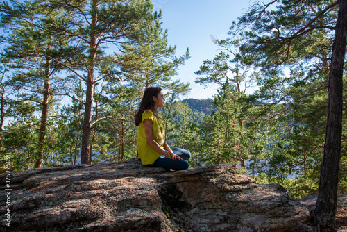 A girl with long hair is sitting on a rock in the forest against the background of a lake and pines.