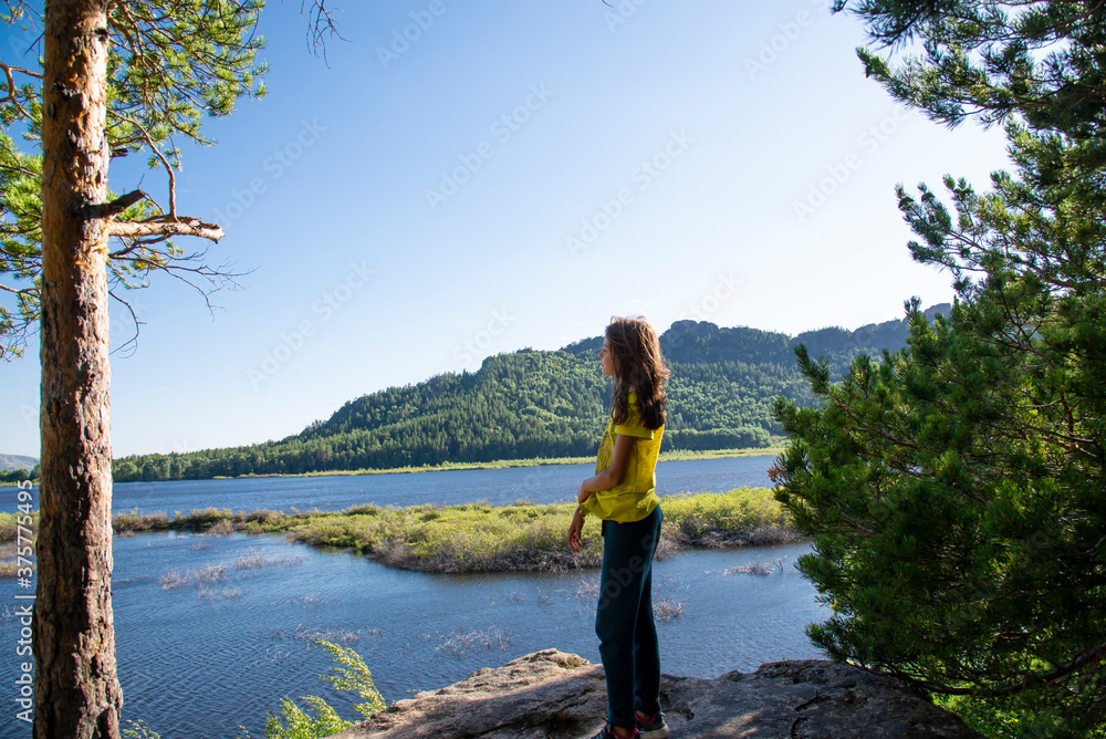 A girl with long hair stands on a rock cliff with pine trees and looks at the lake.