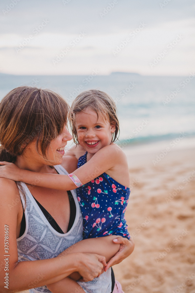 Young mom smiling and holding her daughter on tropical beach at sunset Young mom smiling and holding her daughter on tropical beach at sunset