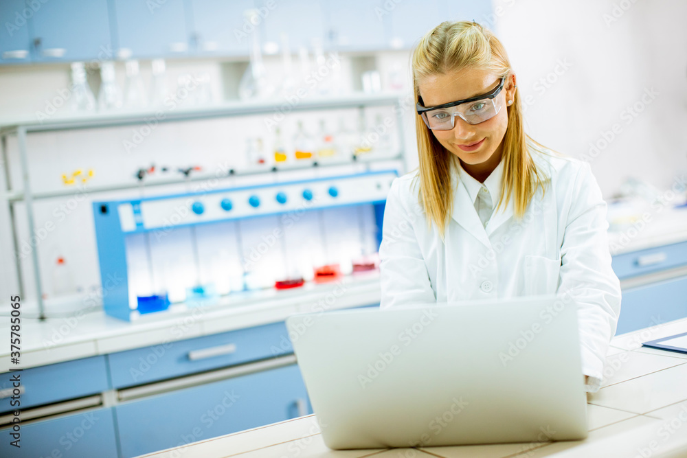 Female researcher in white lab coat and protective goggles using laptop while working in the laboratory Female researcher in white lab coat and protective goggles using laptop while working in the laboratory