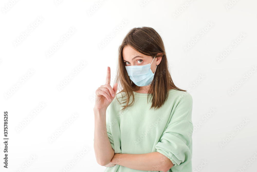 Serious female wearing medical mask during coronavirus standing on white background at home and pointing up with index finger while looking at camera Serious female wearing medical mask during coronavirus standing on white background at home and pointing up with index finger while looking at camera