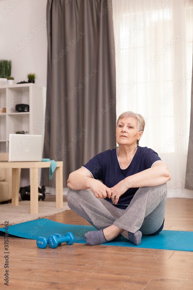 Woman sitting on yoga mat waiting for the wellness trainer. Old person pensioner online internet exercise training at home sport activity with dumbbell, resistance band, swiss ball at elderly