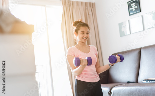 A woman wearing sports clothes raised a dumbbell exercise in the living room. She is training at home. woman in sportswear with Purple dumbbells in her hands.
