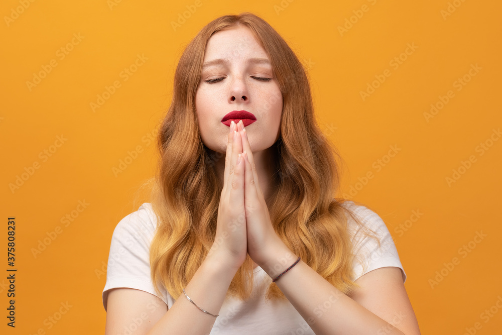 Pretty girl with wavy redhead, holding palms together, praying for peace and love, having peaceful facial expression Pretty girl with wavy redhead, holding palms together, praying for peace and love, having peaceful facial expression