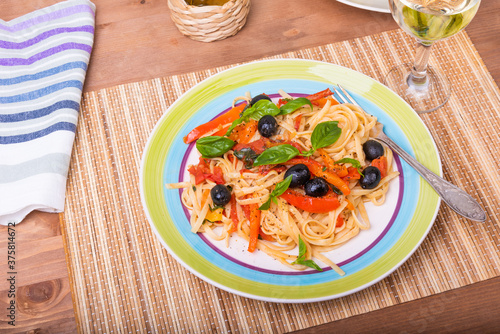 Vegetarian pasta with vegetables, linguine with bell pepper, tomatoes and olives on a plate standing on a napkin on the table and a glass of dry white wine, close-up
