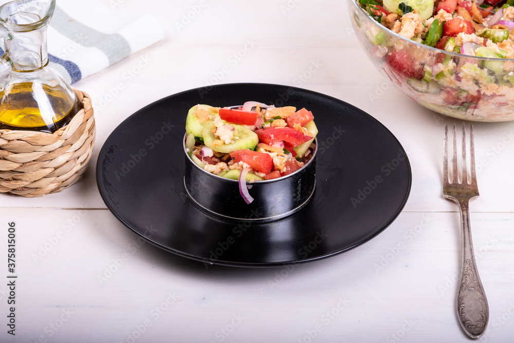 Italian Tuscan panzanella salad, mixed salad of bread and vegetables in a culinary ring on a black plate and a glass bowl with ready-made salad on a wooden light table, close-up Italian Tuscan panzanella salad, mixed salad of bread and vegetables in a culinary ring on a black plate and a glass bowl with ready-made salad on a wooden light table, close-up