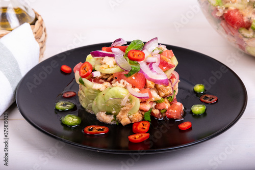 Italian tuscan salad panzanella, mixed salad of bread and vegetables on a black plate decorated with chilli peppers and a glass bowl with ready-made salad on a wooden light table, close-up