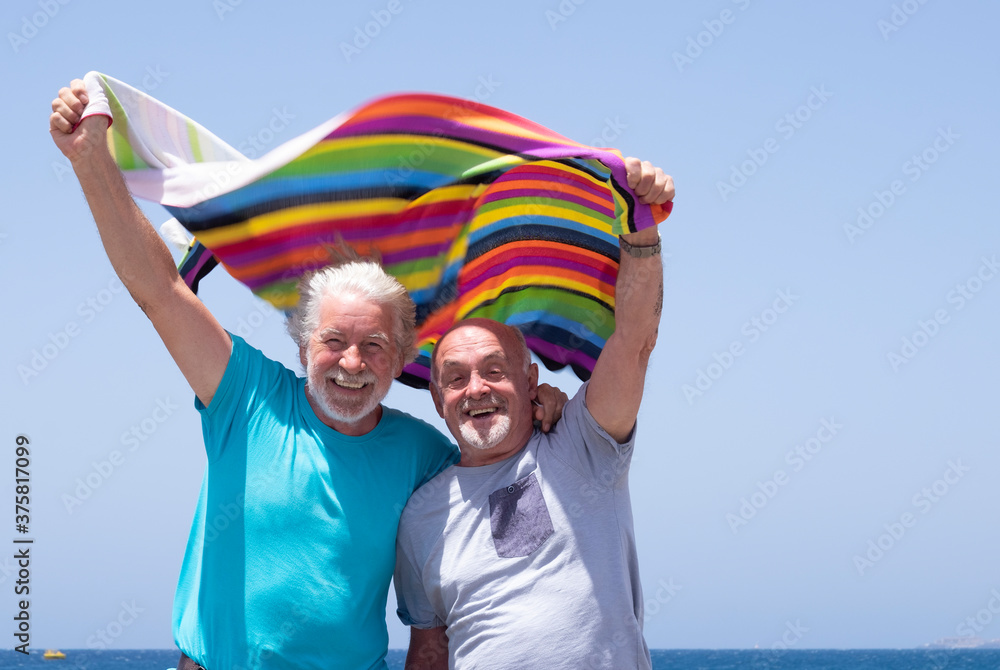 Cheerful couple of senior bearded brothers laughing at the beach holding a colorful towel in the wind - concept of freedom and happiness in sea holidays