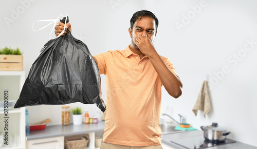 recycling, sorting and sustainability concept - young indian man holding stinky trash bag over home kitchen background