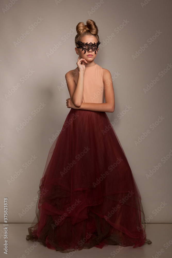 Beautiful little girl child in a lace mask and a ball gown with an evening hairstyle in the studio on a gray background