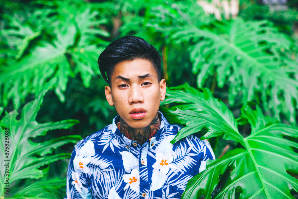 Young asian tattooed man standing among palm tree leaves. Young asian tattooed man standing among palm tree leaves.