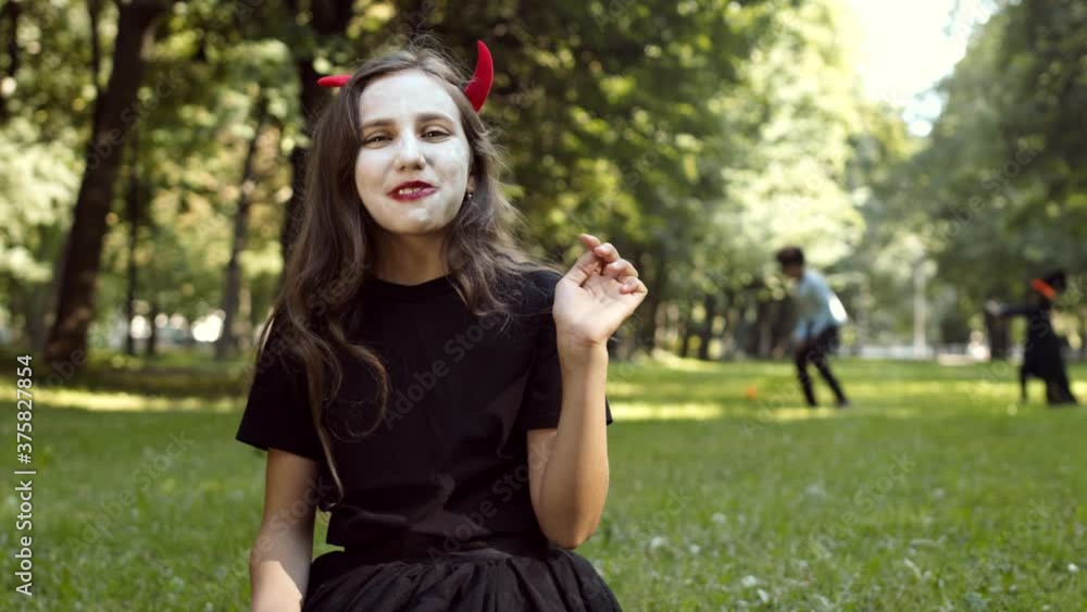 Cheerful Caucasian girl wearing devil costume is sitting on grass in park, taking sweets out of pumpkin buckets and eating them while other kids playing on background Cheerful Caucasian girl wearing devil costume is sitting on grass in park, taking sweets out of pumpkin buckets and eating them while other kids playing on background