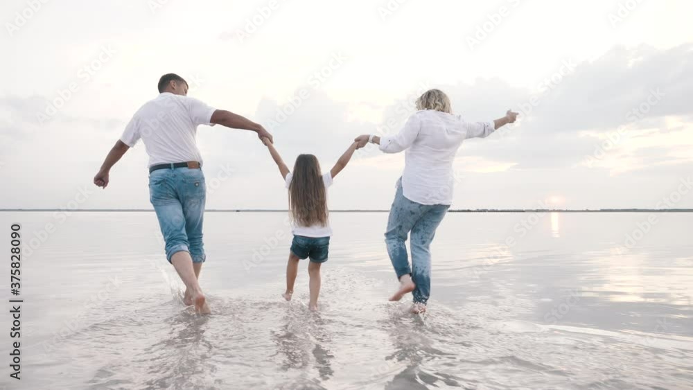 Rear view at active couple carrying little daughter, running on lake water together holding hands. Happy loving family plays funny flying adventure games outdoor, carefree relax joy on summer vacation Rear view at active couple carrying little daughter, running on lake water together holding hands. Happy loving family plays funny flying adventure games outdoor, carefree relax joy on summer vacation