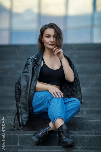 A girl in blue jeans, a black top and a jacket is sitting on the stairs. Portrait of a model in a leather jacket. Urban style.