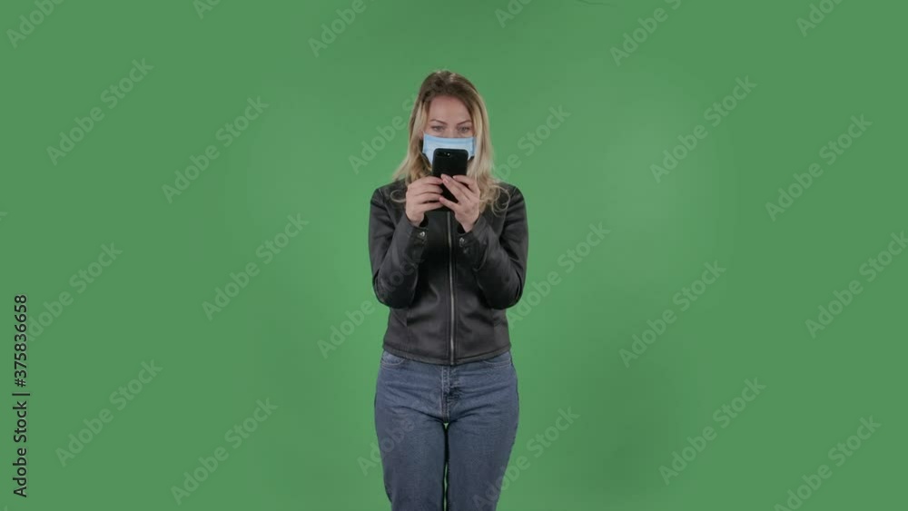 Portrait of beautiful young woman in medical mask is texting on her phone. Blonde with loose hair in a black jacket and jeans on a green screen in the studio. Health Protection Corona Virus Concept. Portrait of beautiful young woman in medical mask is texting on her phone. Blonde with loose hair in a black jacket and jeans on a green screen in the studio. Health Protection Corona Virus Concept.