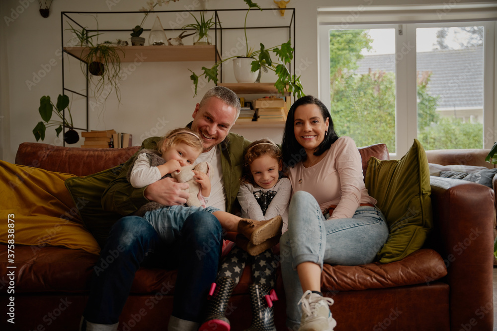 Cheerful couple sitting on couch with two beautiful little daughters at home - one big happy family portrait on a modern couch at home