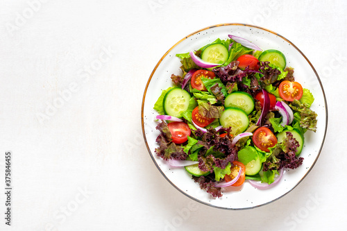Healthy vegetable salad of red cherry tomatoes, cucumber slices, green and purple lettuce leaves, onions and olive oil in plate on white table Top view Flat lay Diet, mediterranean menu Vegan food