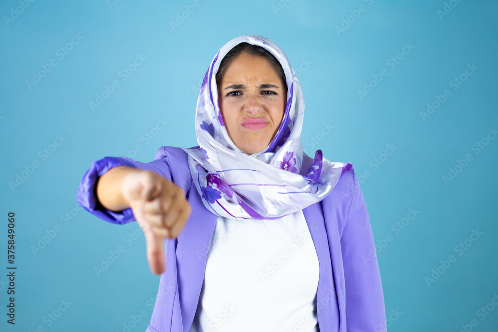 Young beautiful arab woman wearing islamic hijab over isolated blue background with angry face, negative sign showing dislike with thumb down Young beautiful arab woman wearing islamic hijab over isolated blue background with angry face, negative sign showing dislike with thumb down
