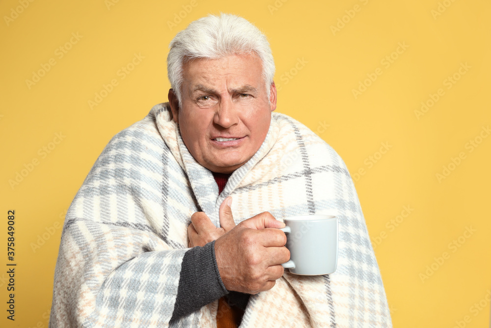 Senior man wrapped in blanket with cup of hot drink on yellow background. Cold symptoms Senior man wrapped in blanket with cup of hot drink on yellow background. Cold symptoms
