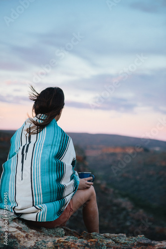 Woman on wrapped in a blue blanket on a mountain top with a cup of coffee overlooking a vallet photo