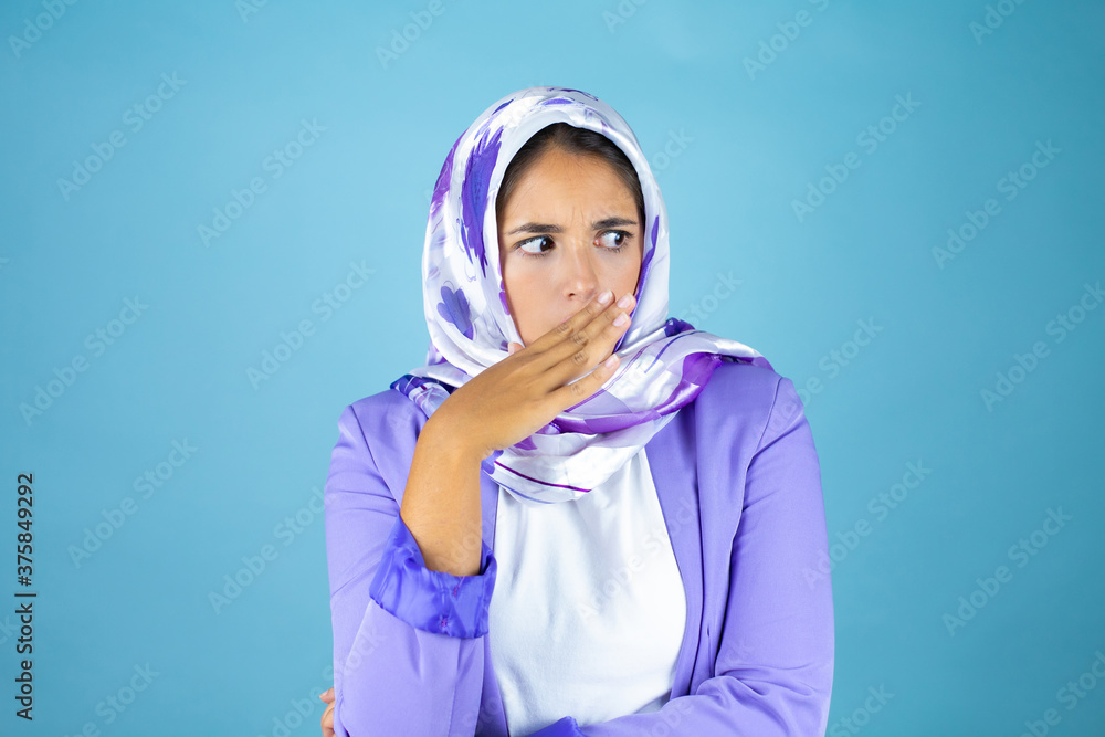 Young beautiful arab woman wearing islamic hijab over isolated blue background surprised covering her mouth Young beautiful arab woman wearing islamic hijab over isolated blue background surprised covering her mouth
