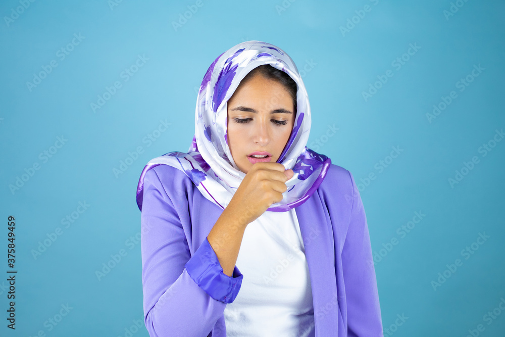 Young beautiful arab woman wearing islamic hijab over isolated blue background with her hand to her mouth because she's coughing Young beautiful arab woman wearing islamic hijab over isolated blue background with her hand to her mouth because she's coughing