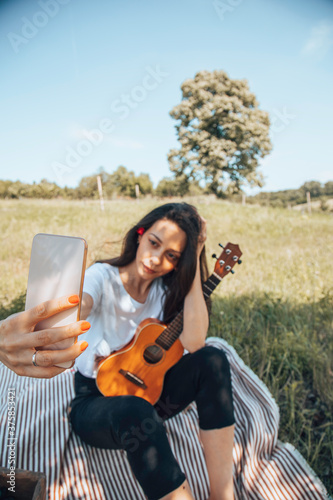 Attractive woman using a cellphone while playing ukulele and enjoying a picnic in nature.
