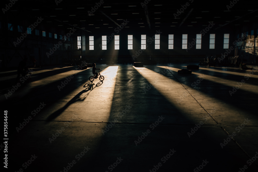 Young child rides a bike through a beam of light, silhouette and shadow Young child rides a bike through a beam of light, silhouette and shadow