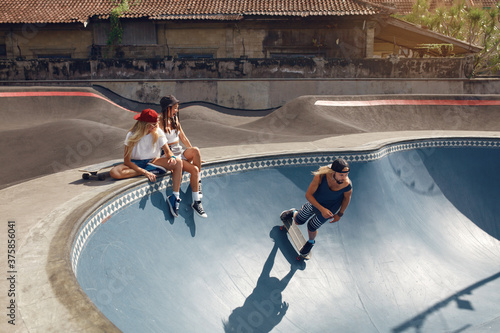 Summer. Skater Friends With Skateboard At Skatepark. Group Of People In Casual Outfit Outdoor. Hipster Girls Sitting On Concrete Ramp, Guy Skateboarding. Extreme Sport As Urban Lifestyle.