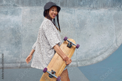 Portrait Of Skater Girl In Skatepark. Female Teenager In Casual Outfit With Skateboard Against Concrete Wall. Summer Skateboarding With Modern Urban Sport Equipment As Part Of Active Lifestyle. photo