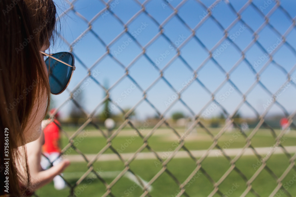 Teenage girl wearing sunglasses watches baseball game through fence Teenage girl wearing sunglasses watches baseball game through fence