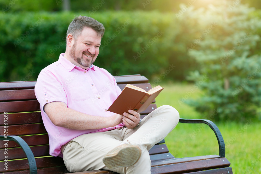 An adult man with a short beard in summer clothes is reading a book while sitting on a bench in the park An adult man with a short beard in summer clothes is reading a book while sitting on a bench in the park