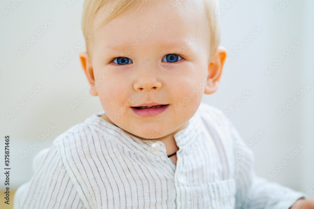Baby boy with blue eyes and blonde hair wearing striped shirt smiling at camera, light background. Closeup portrait of cheerful child indoors. Concept of babyhood Baby boy with blue eyes and blonde hair wearing striped shirt smiling at camera, light background. Closeup portrait of cheerful child indoors. Concept of babyhood
