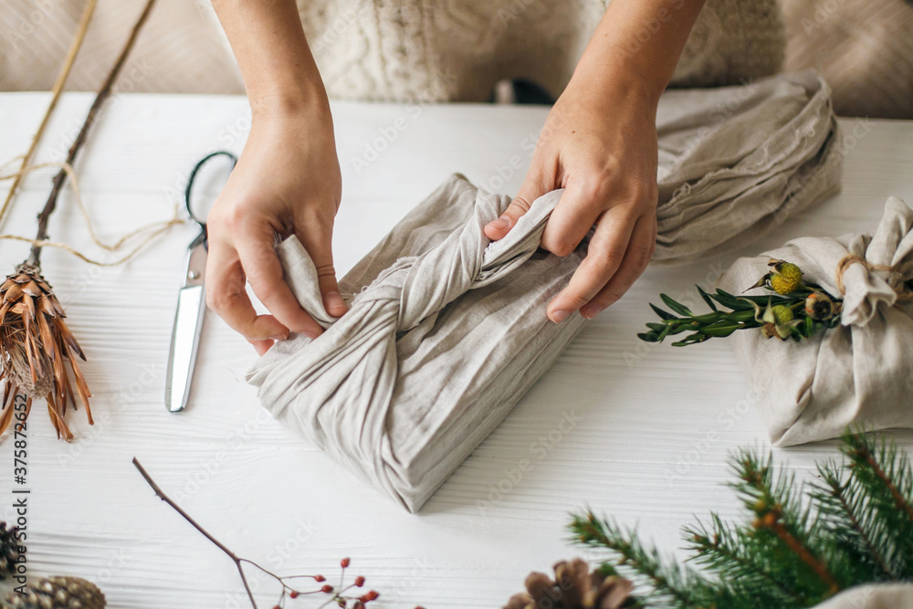 Hands wrapping christmas gift in linen fabric in furoshiki style on white  rustic table with natural fir, herb, pine cones and scissors. Female  preparing plastic free christmas present Stock Photo | Adobe