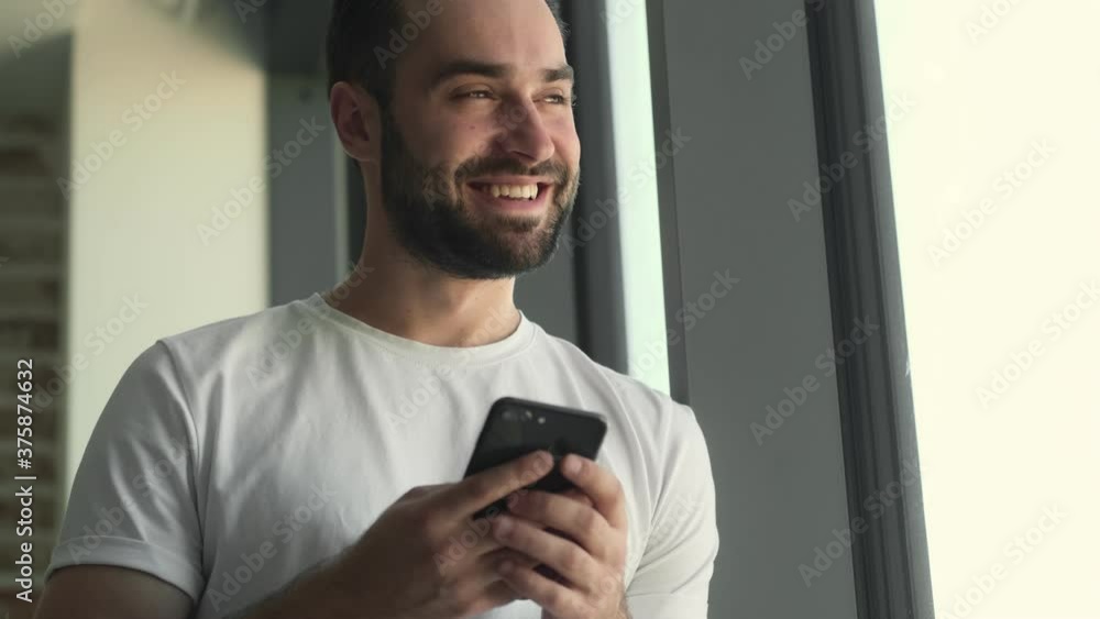 Young happy man standing near window indoors at home while using mobile phone Young happy man standing near window indoors at home while using mobile phone