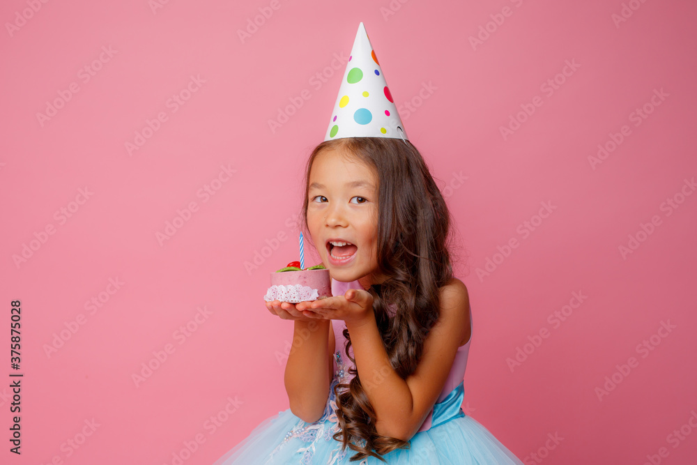 little asian girl celebrates birthday by holding a cake with a candle to bite the cake on a pink background