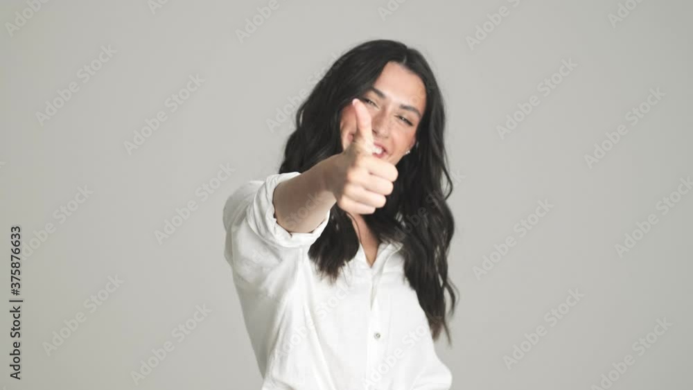 A smiling young woman is pointing to the viewer and showing thumb-up gesture standing isolated over gray background A smiling young woman is pointing to the viewer and showing thumb-up gesture standing isolated over gray background