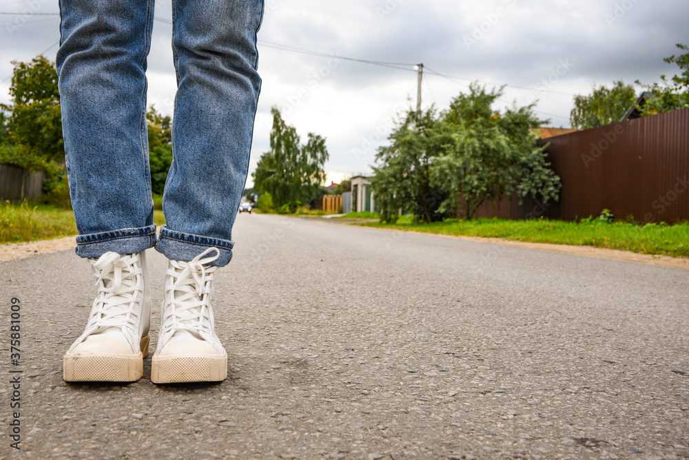 Lifestyle. Modern young woman in a stylish blue jeans in fashionable white sneakers stands on road. Stylish women's shoes. Youth style. Closeup of female legs in a trendy shoes. Lifestyle. Modern young woman in a stylish blue jeans in fashionable white sneakers stands on road. Stylish women's shoes. Youth style. Closeup of female legs in a trendy shoes.