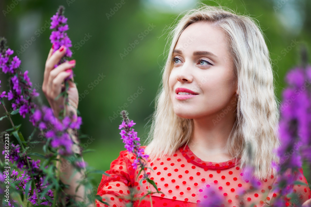 Young woman with flowers in the park. Smiling blonde in a red dress enjoys the scent. Young woman with flowers in the park. Smiling blonde in a red dress enjoys the scent.