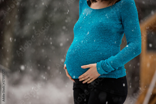 Pregnant woman in blue sweater standing outside on a snowy day touching her swollen belly photo