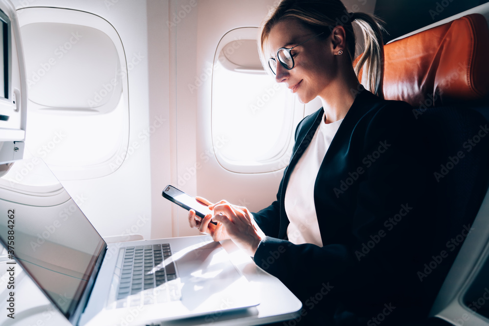 Joyful adult entrepreneur using smartphone while working on laptop in plane Joyful adult entrepreneur using smartphone while working on laptop in plane