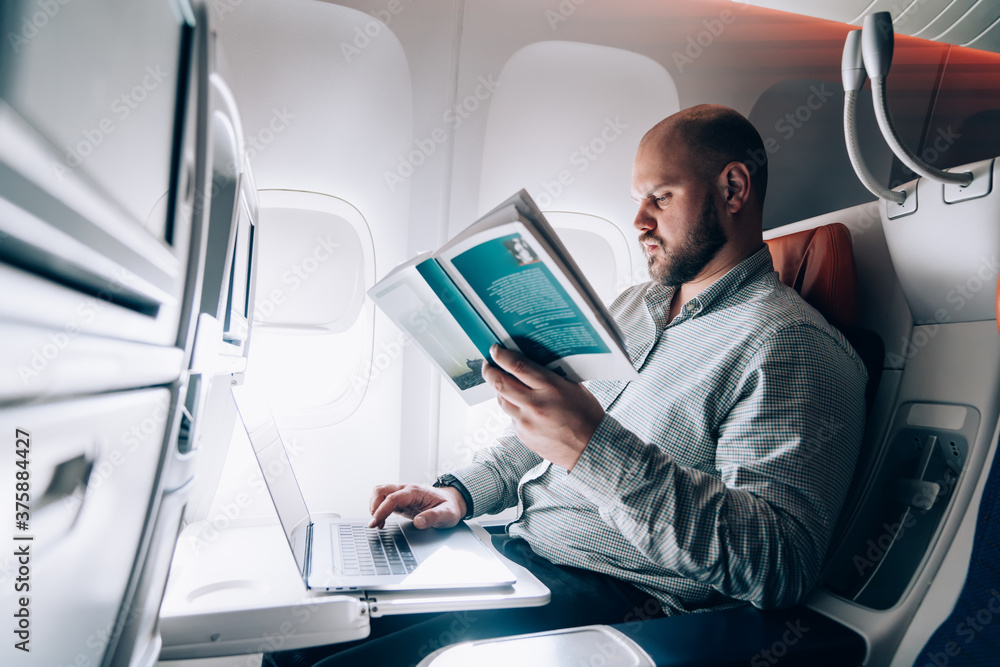 Fototapeta premium Serious man typing on laptop and reading book in aircraft