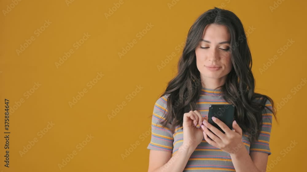 A smiling young woman is nodding while using her smartphone standing isolated over yellow background A smiling young woman is nodding while using her smartphone standing isolated over yellow background