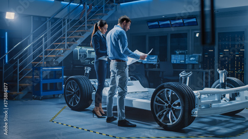 Auto Industry Design Facility: Male Chief Engineer Shows Car Blueprints Female Software Design and Integration Engineer. Electric Vehicle Platform Chassis Concept Has Wheels, Engine and Battery  © Gorodenkoff