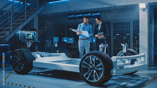 Auto Industry Design Facility: Male Chief Engineer Shows Car Blueprints Female Software Design and Integration Engineer. Electric Vehicle Platform Chassis Concept Has Wheels, Engine and Battery 