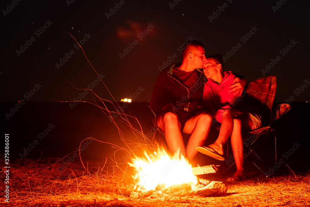 Happy Young couple sitting by the fire. Big bonfire and young couple in the background. Long exposure photo. Happy Young couple sitting by the fire. Big bonfire and young couple in the background. Long exposure photo.